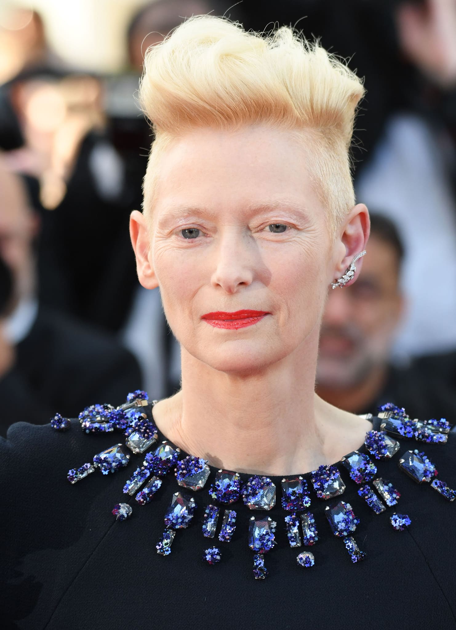 Blue crystals and sequins frame Swinton’s neckline like jewelry, complemented by her sculpted blonde undercut, red lipstick, and diamond ear cuff