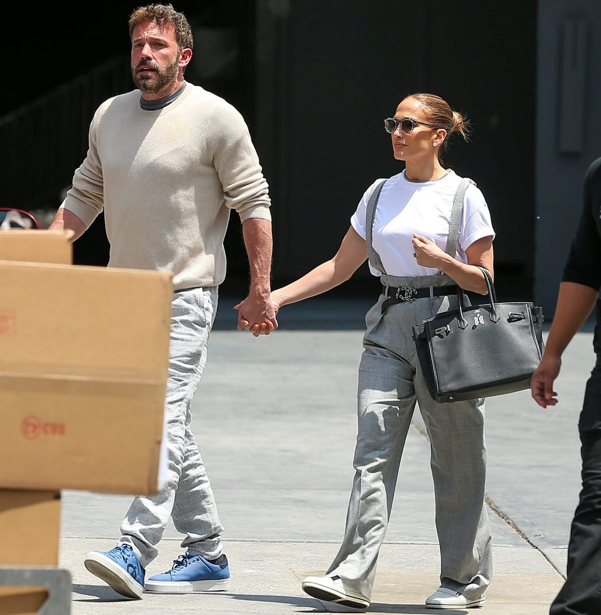 Ben Affleck and Jennifer Lopez hold hands outside RED Studios in Hollywood on May 3, 2022, with Jennifer showcasing “quiet luxury” in a Brunello Cucinelli gray wool jumpsuit, a cropped tee from The Great, Valentino Rockstud sunglasses, and Roger Vivier “Sneaky Viv” zip-up sneakers, paired with her signature Hermès Birkin bag