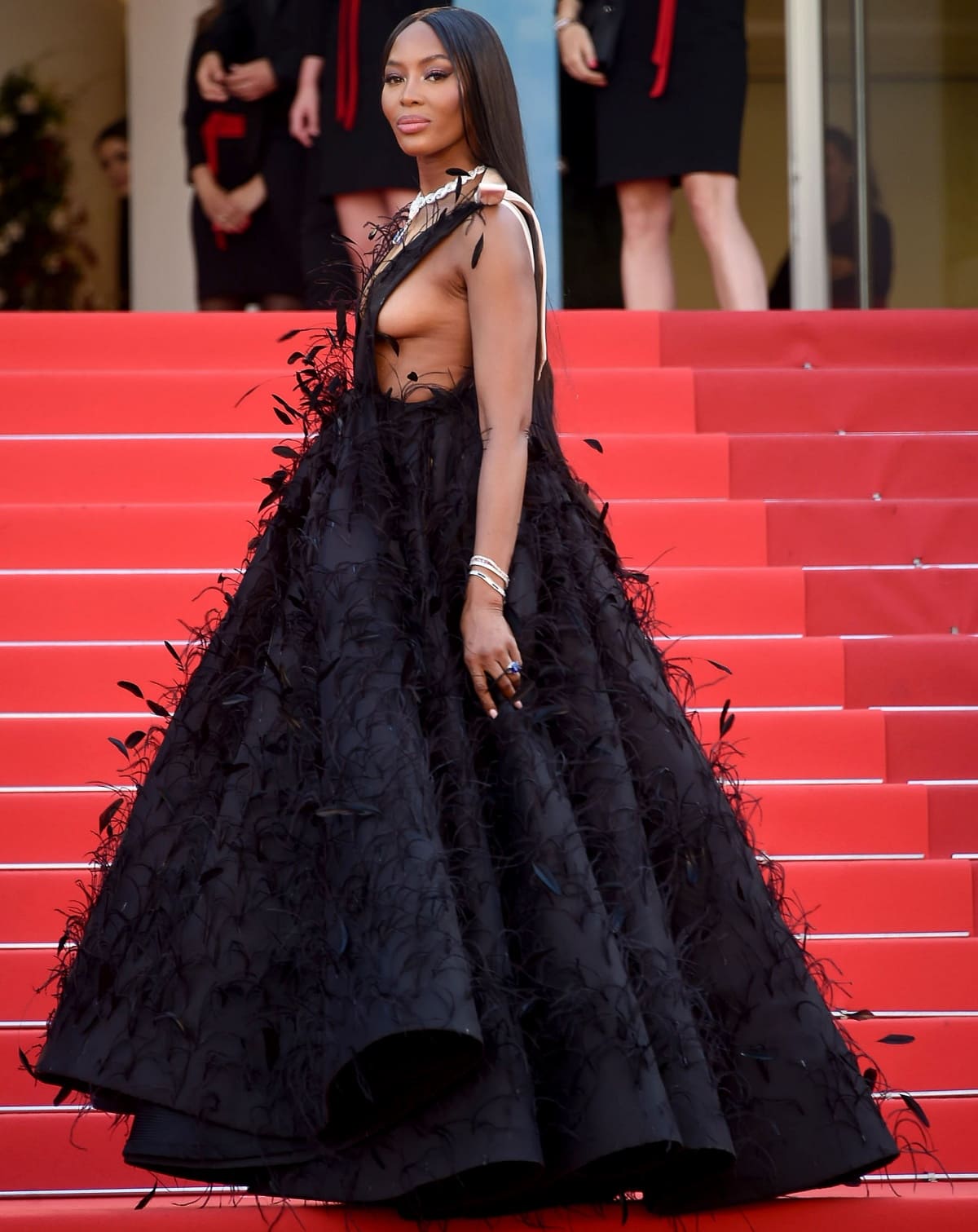 Naomi Campbell commands the Cannes red carpet in a black Valentino Spring 2022 Haute Couture gown featuring a plunging bodice, feather embellishments, and a voluminous floor-sweeping skirt