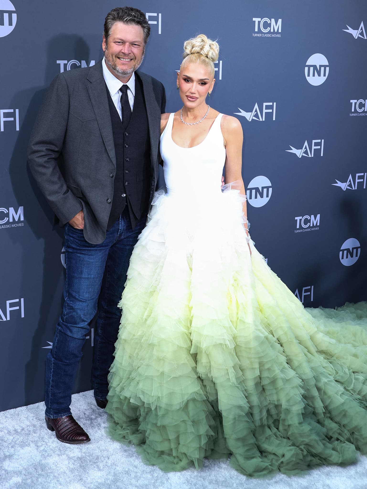 Blake Shelton keeps it classic in a charcoal blazer, waistcoat, and jeans with brown leather boots as he joins Gwen Stefani—stunning in a white-to-green ombré Nicole + Felicia couture gown—on the red carpet at the 48th AFI Life Achievement Award Gala Tribute on June 9, 2022