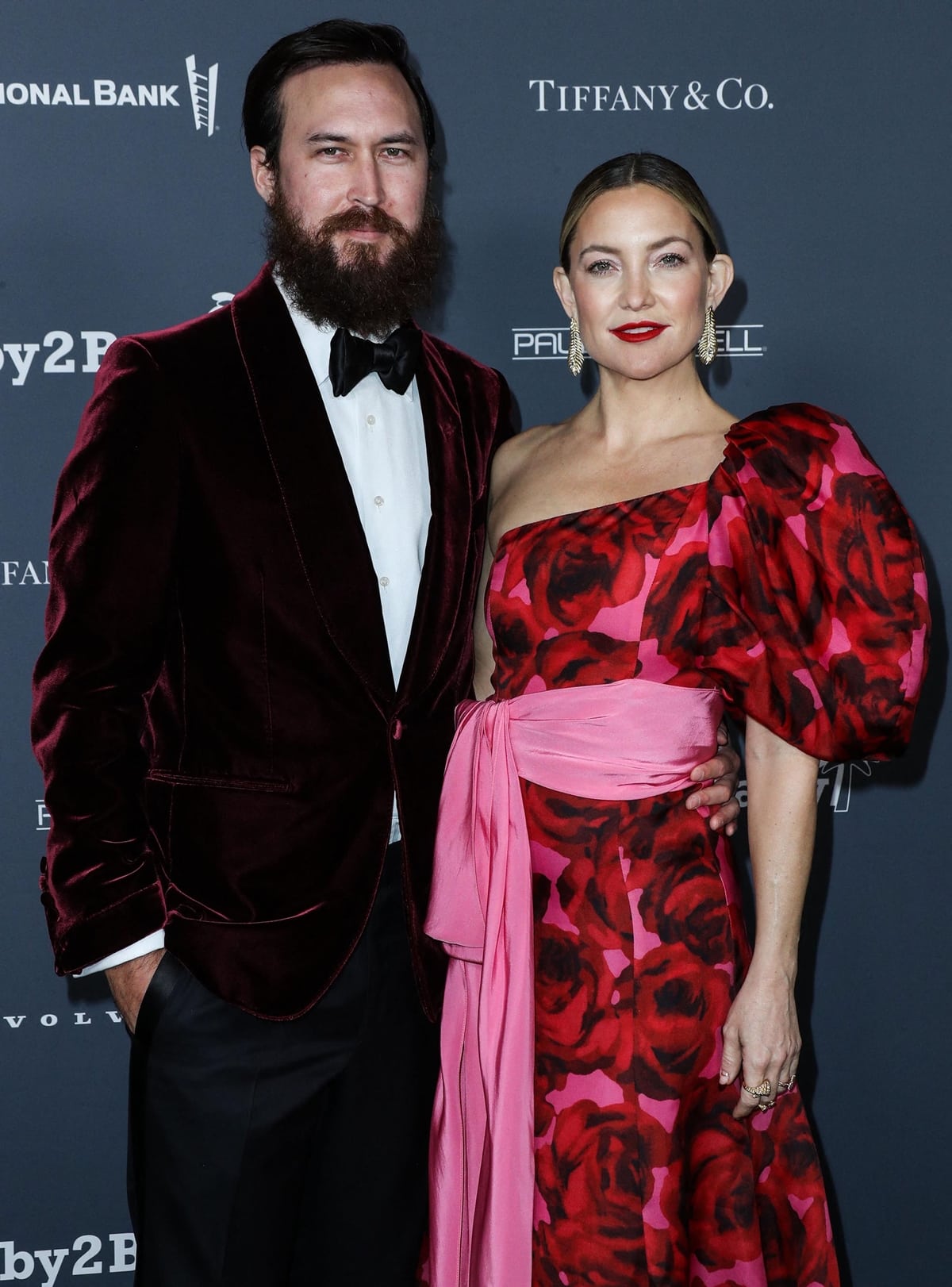 Kate Hudson stuns in a red and pink floral gown with a bold puff sleeve and waist-tied sash, standing next to fiancé Danny Fujikawa at a red carpet event