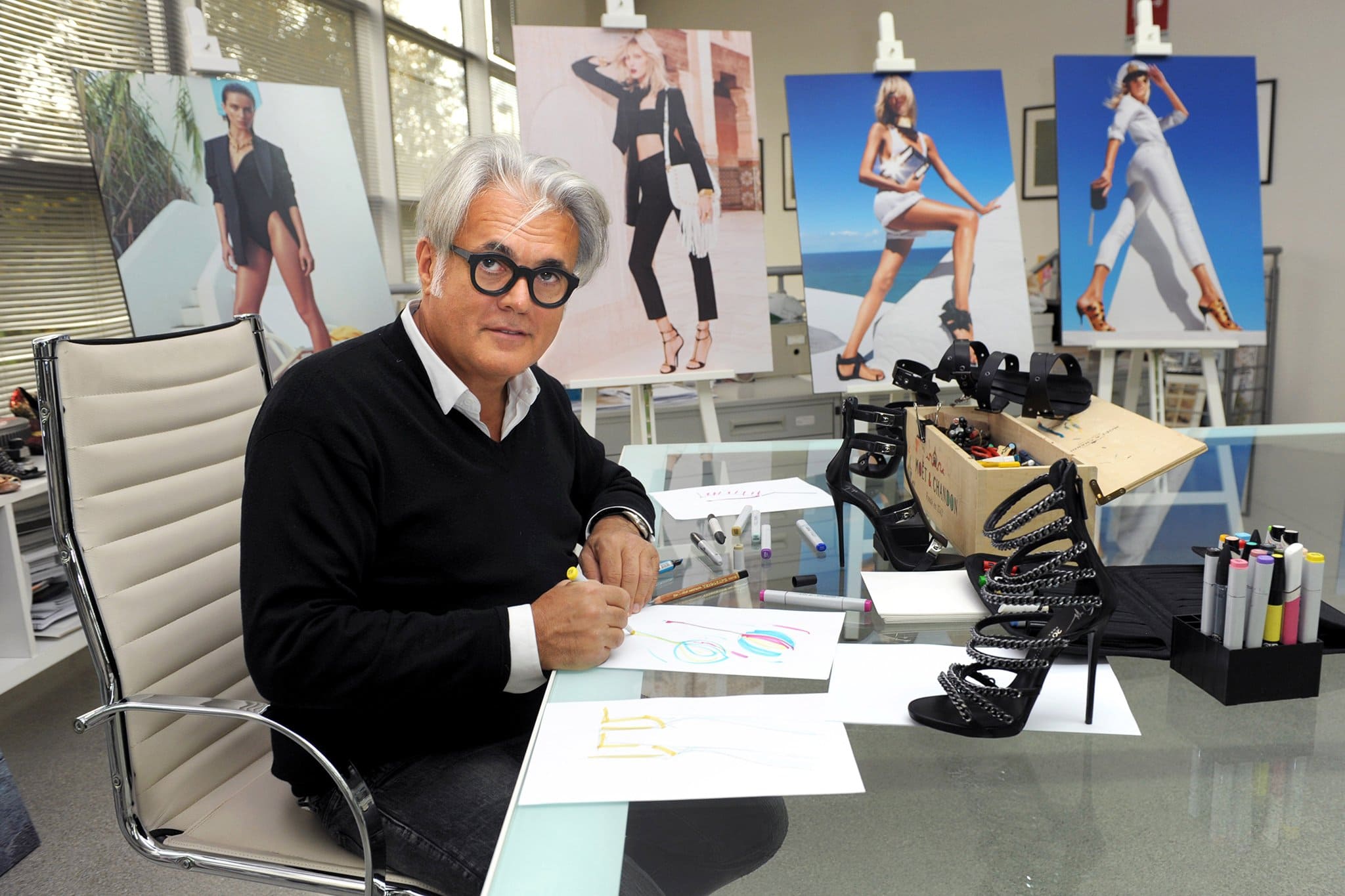 Giuseppe Zanotti, in his design studio, sketches one of his signature creations surrounded by prototypes and striking campaign imagery, showcasing his artistry and dedication to luxury footwear
