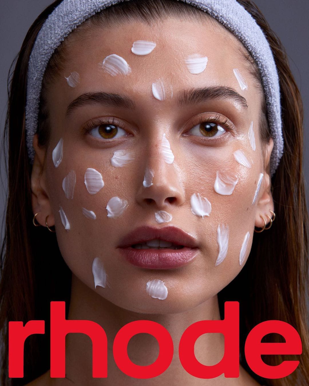 Hailey Bieber appears in Rhode’s launch campaign on June 15, 2022, wearing a plush spa-style headband while showcasing the skincare brand’s minimalist, dewy aesthetic with dotted moisturizer applied across her face beneath Rhode’s bold signature logo