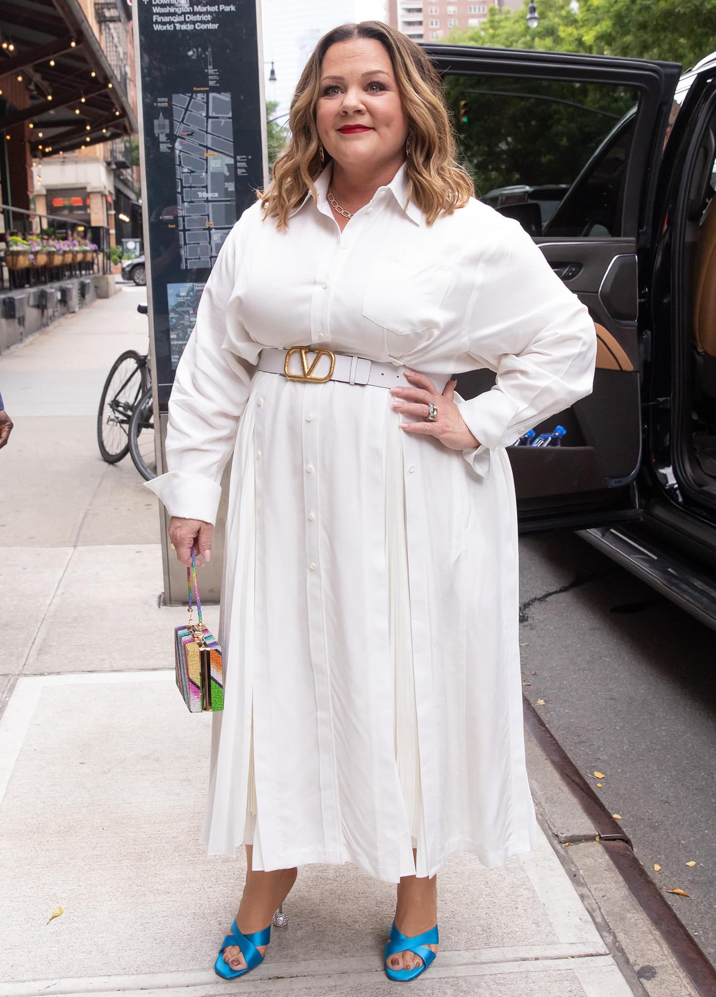 On June 7, 2022, Melissa McCarthy stepped out in New York City wearing a flowing white Valentino shirtdress cinched with a V-logo belt, elevating the monochrome look with vibrant blue Aquazzura “Yes Darling” satin mules featuring crystal disco-ball heels as she promoted her new shows on The Tonight Show Starring Jimmy Fallon