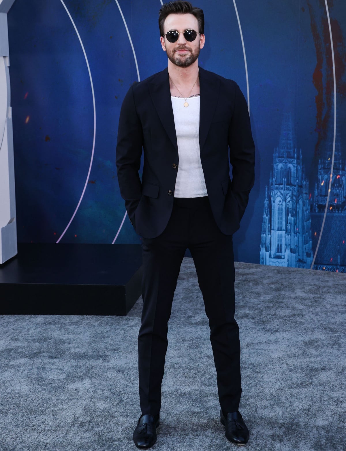 Chris Evans keeps the focus on understated tailoring in a navy Eleventy Milano suit, pairing the sharp silhouette with a white ribbed tank, tassel-trimmed black loafers, and round Ray-Ban aviators