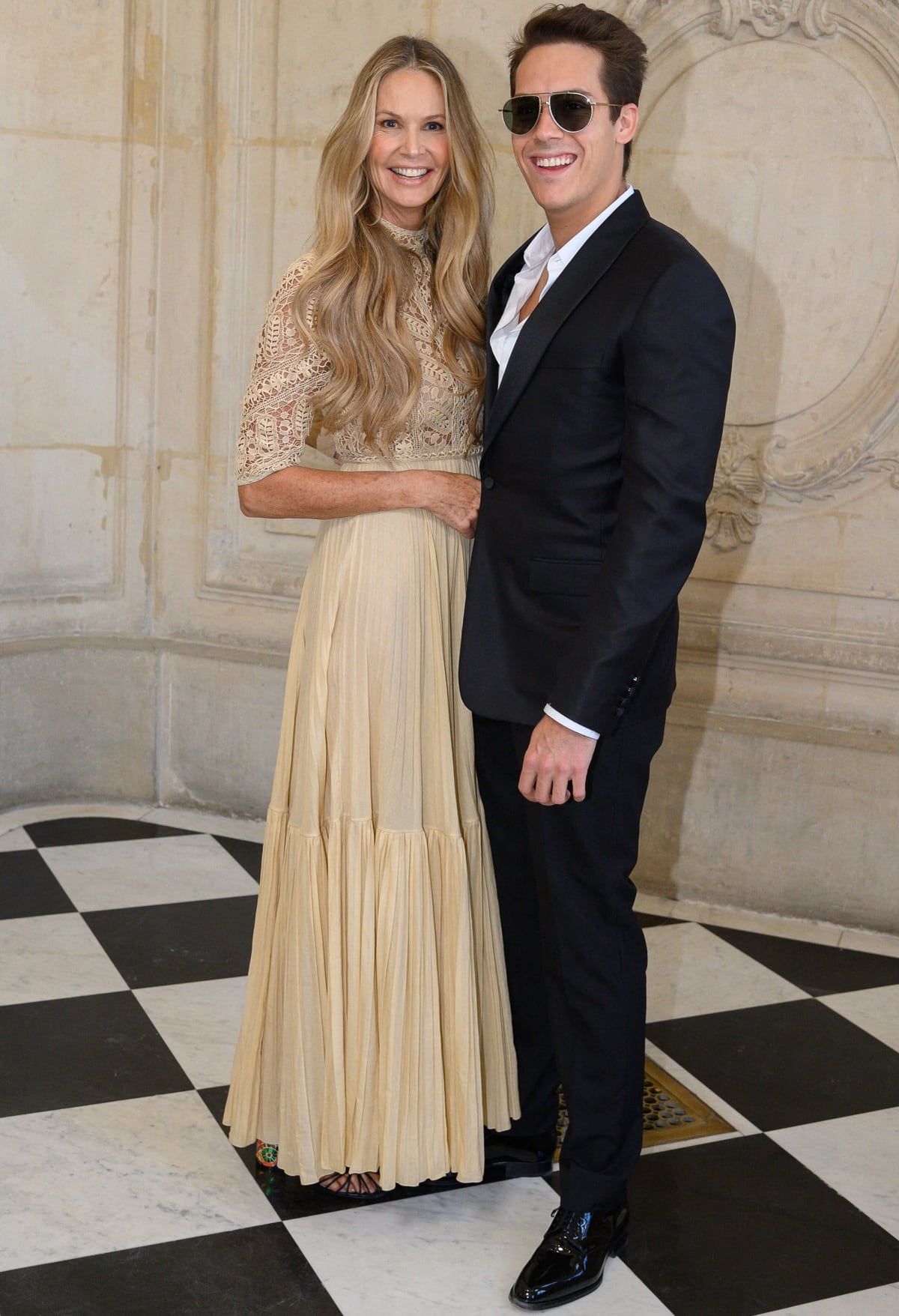 Elle MacPherson glows in a beige lace Dior dress paired with colorful platform heels, posing alongside her son Flynn Busson at the Dior Haute Couture show