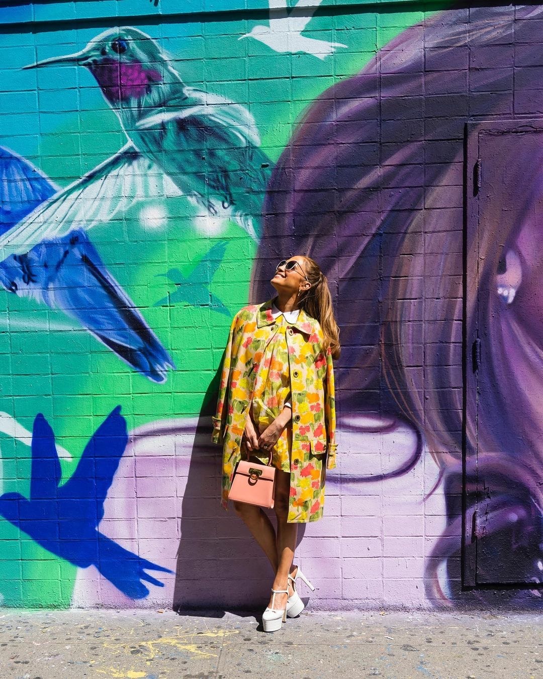 Jennifer Lopez poses in front of a vibrant New York mural wearing a floral Coach coat and towering white Gucci platform pumps that amplified the playful retro mood of the look