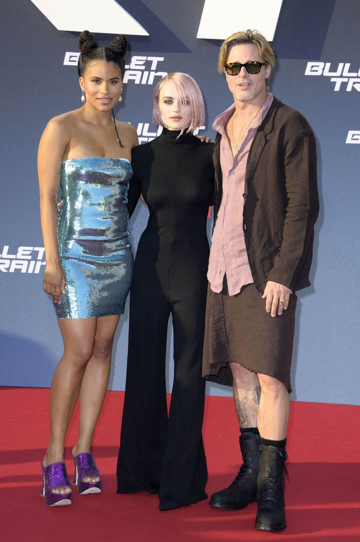 Brad Pitt poses with co-stars Zazie Beetz and Joey King at the Bullet Train Berlin premiere, where Beetz turned heads in a shimmering blue sequined mini dress with bold purple platform mules, King embraced futuristic chic in a sleek black Balenciaga jumpsuit, and Pitt made headlines in his effortlessly cool brown linen skirt set