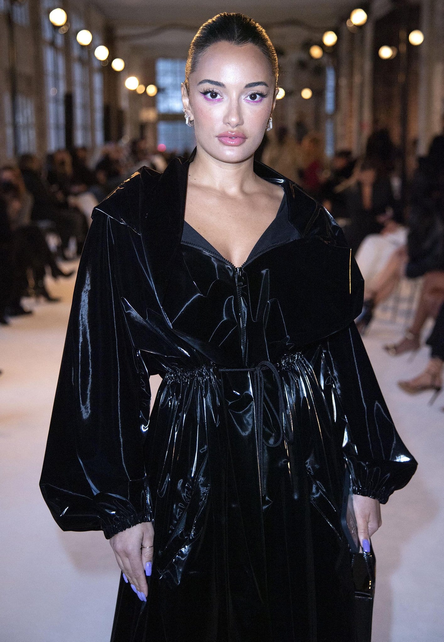 Amina Muaddi attends a fashion show in Paris, turning heads in a sleek black patent-look coat with bold makeup and minimalist accessories