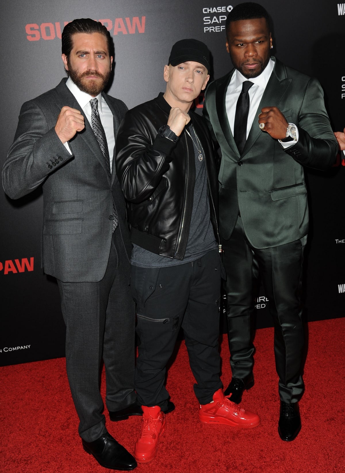 Eminem stands between the much taller Jake Gyllenhaal and 50 Cent at the Southpaw premiere in New York on July 20, 2015, rocking a black leather jacket, cargo pants, and eye-catching red Air Jordan 4 sneakers