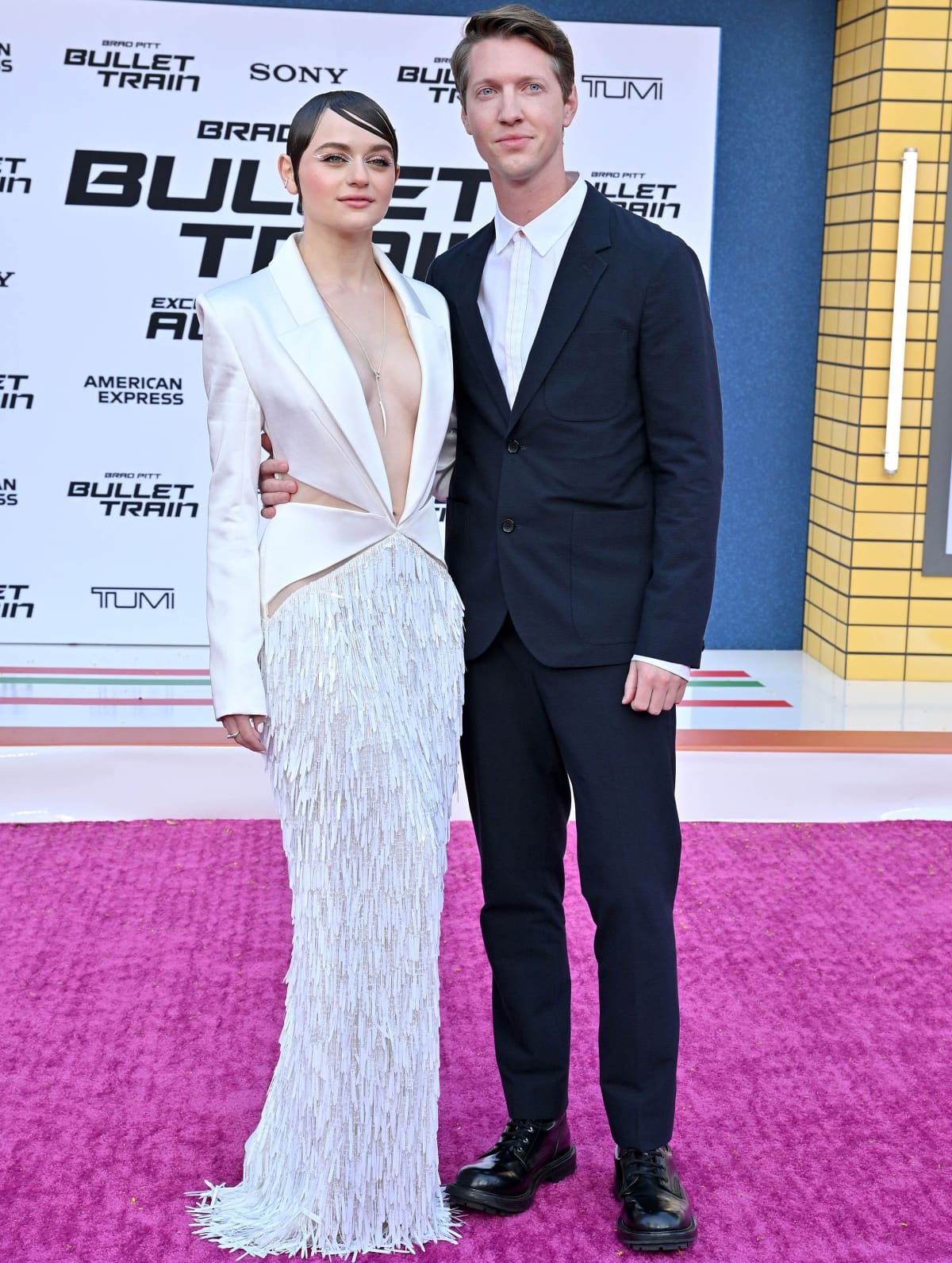Joey King exudes sleek, modern glamour in a plunging white Cong Tri blazer dress with fringe detailing, joined by fiancé Steven Piet in a tailored navy suit at the Bullet Train Los Angeles premiere on August 1, 2022