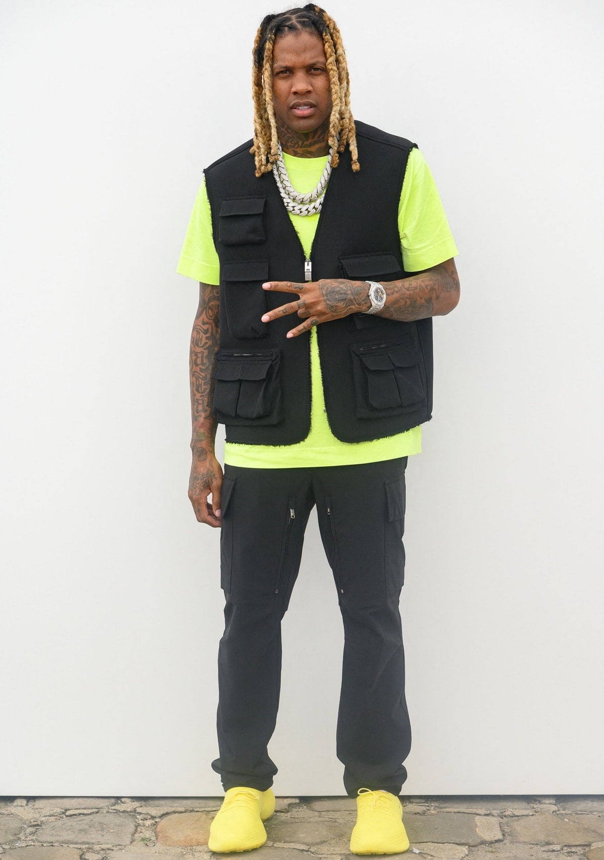 Lil Durk stands out in a neon yellow tee with matching sneakers—possibly Yeezys—paired with a black tactical vest and cargo pants while attending the Givenchy show during Paris Fashion Week on June 22, 2022