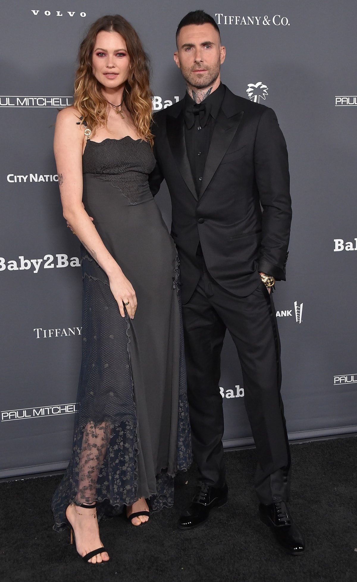Behati Prinsloo, wearing a sheer-accented black gown and strappy heels, appears slightly taller than husband Adam Levine—though she's shorter than him without heels