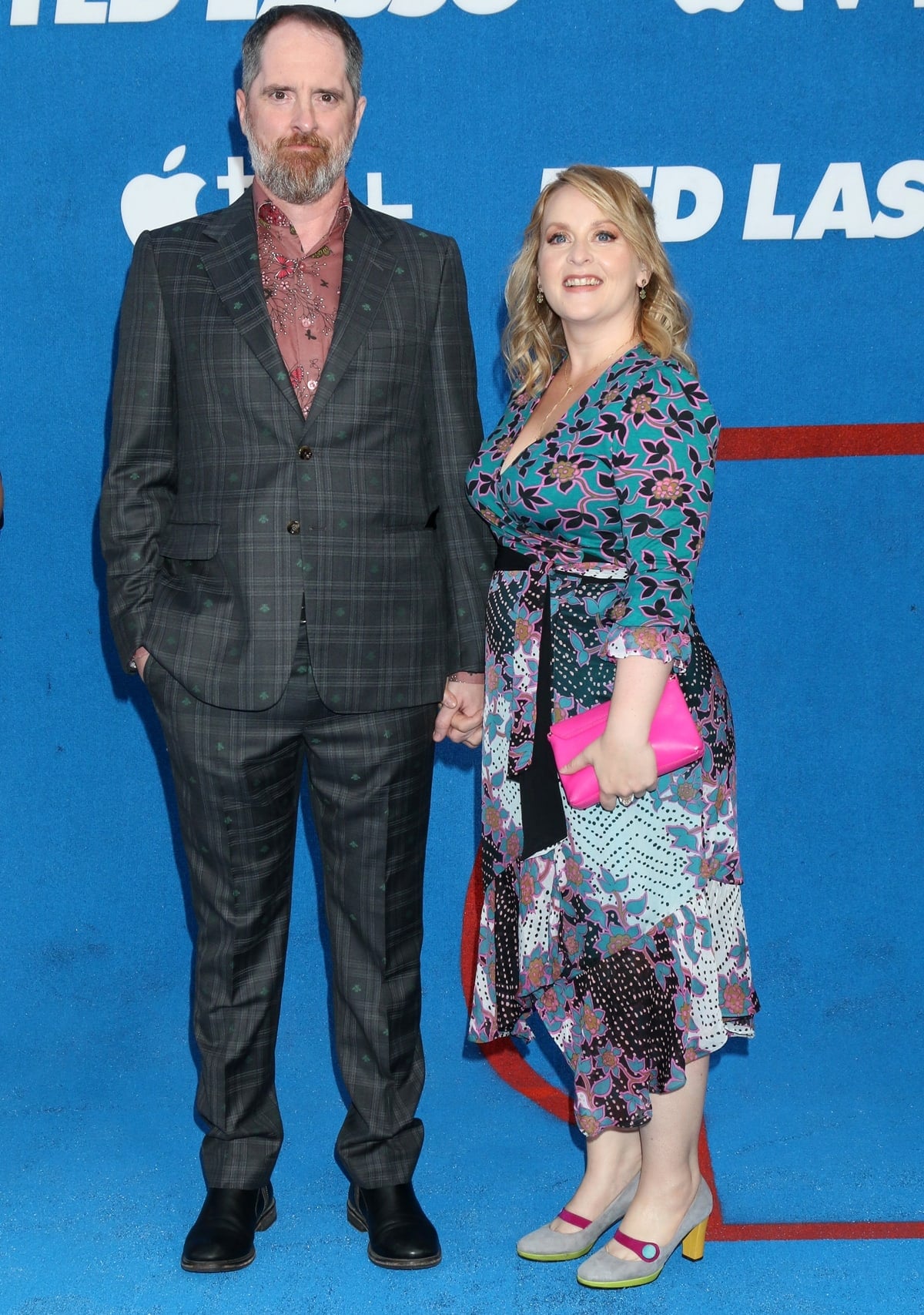 Ted Lasso co-creator Brendan Hunt keeps it quirky in a checkered suit with floral embroidery as he poses with wife Shannon Nelson, who adds a playful pop of color in a floral wrap dress, colorblock heels with yellow soles, and a hot pink clutch