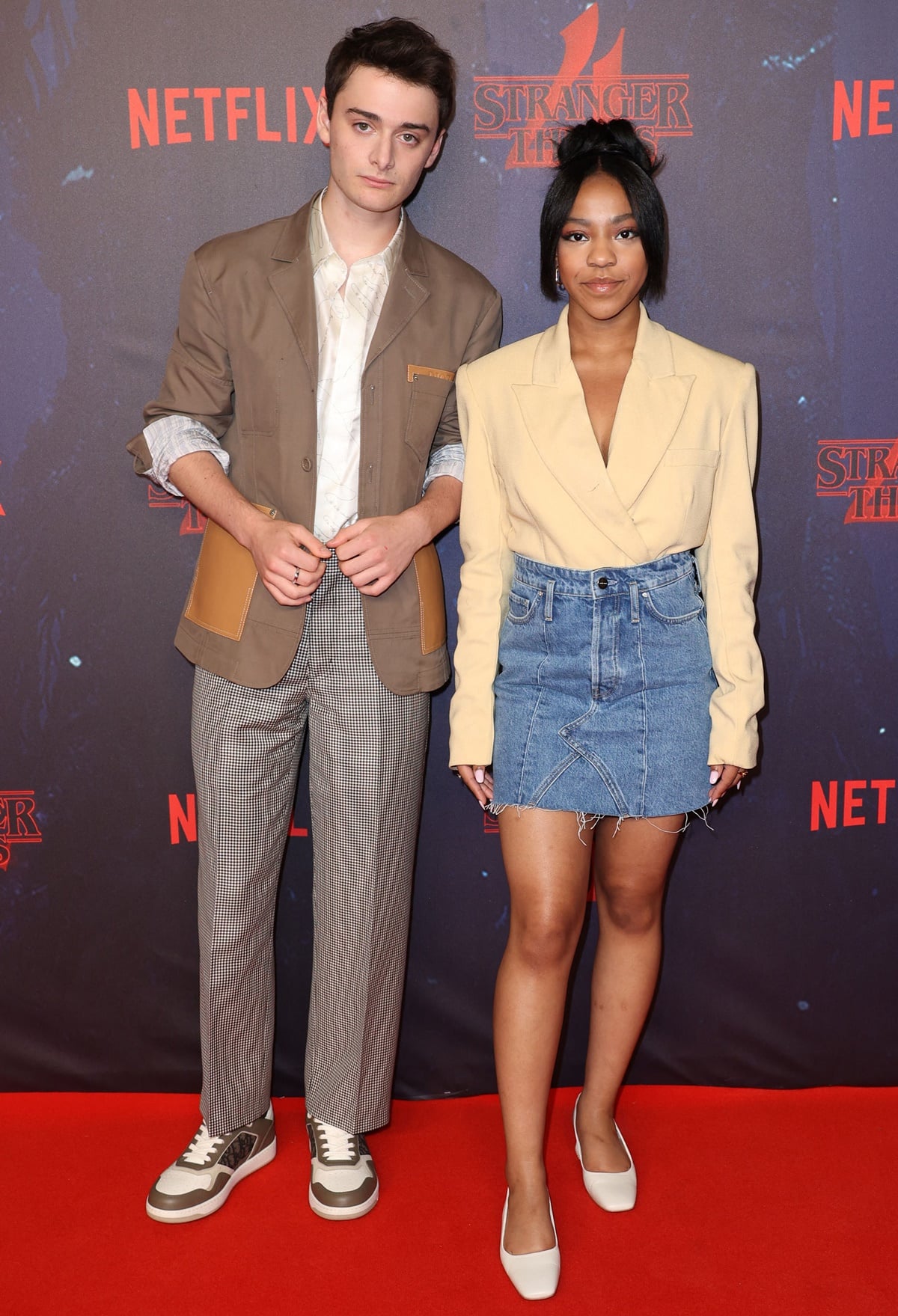 Noah Schnapp kept it casual in checkered pants and colorblock sneakers, while Priah Ferguson added polish to her raw-hem denim mini skirt with cream square-toe heels at the Stranger Things Festival photocall in Paris on May 24, 2022