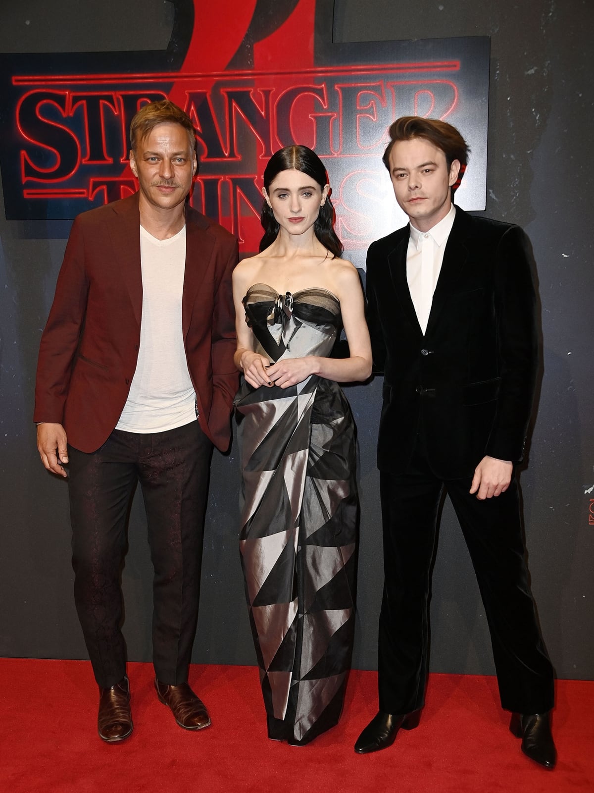 Natalia Dyer turned heads in a sculptural strapless Vivienne Westwood gown featuring a metallic geometric pattern, posing alongside her dapper co-stars Tom Wlaschiha in a burgundy blazer and Charlie Heaton in a sharp black velvet suit at the Stranger Things Season 4 screening in Berlin on May 23, 2022