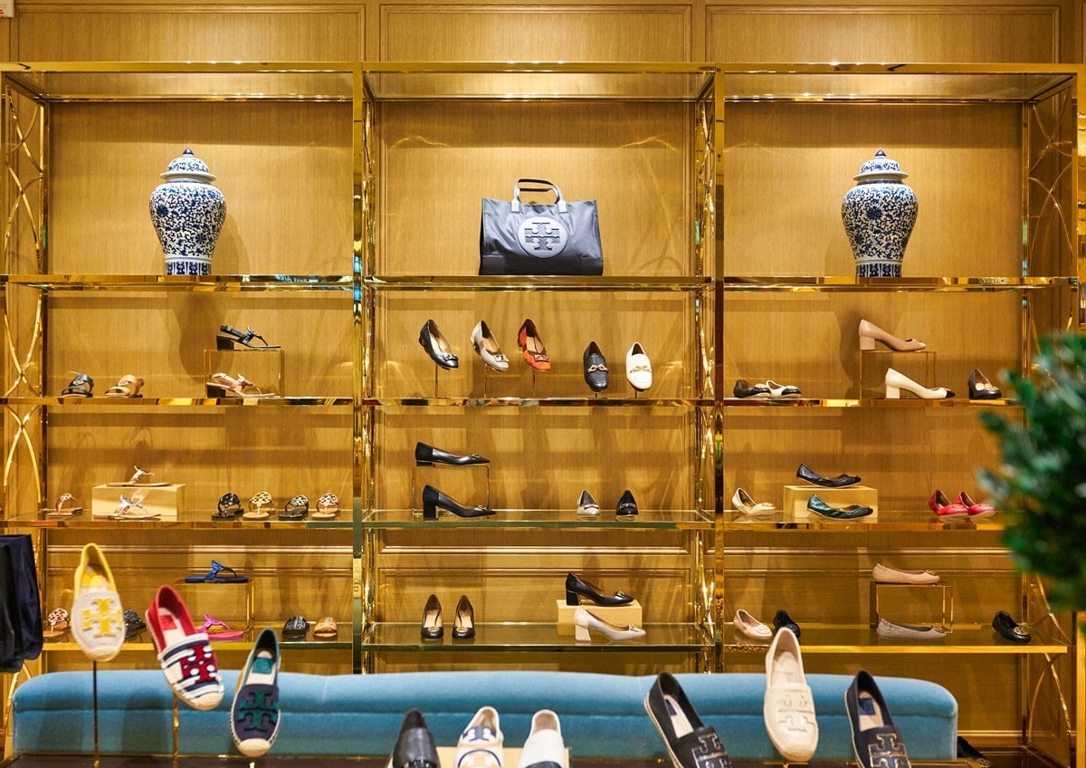 Tory Burch’s official boutique showcases a curated selection of handbags and shoes arranged on gleaming gold shelves that reflect the brand’s polished, attainable-luxury aesthetic