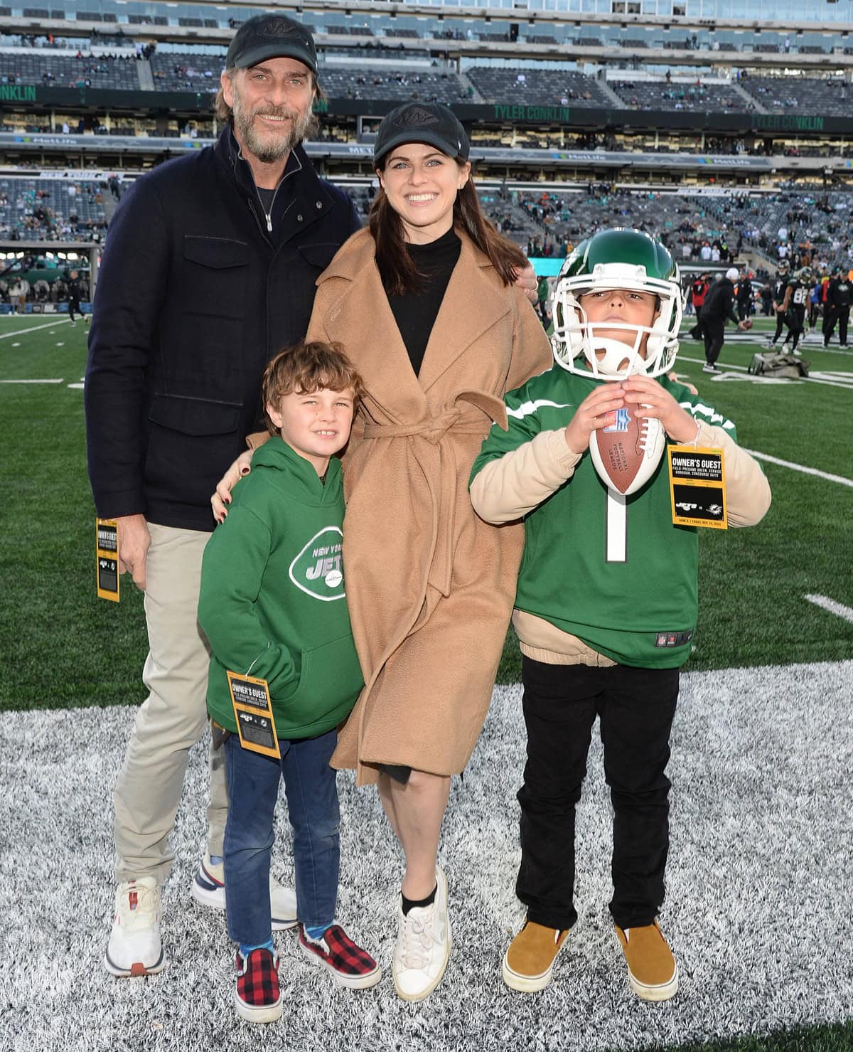 Alexandra Daddario keeps it cozy and casual in white lace-up sneakers and a camel wrap coat as she enjoys a sweet family moment with husband Andrew Form and his sons Rowan and Julian at the Miami Dolphins vs. New York Jets game at MetLife Stadium on November 24, 2023