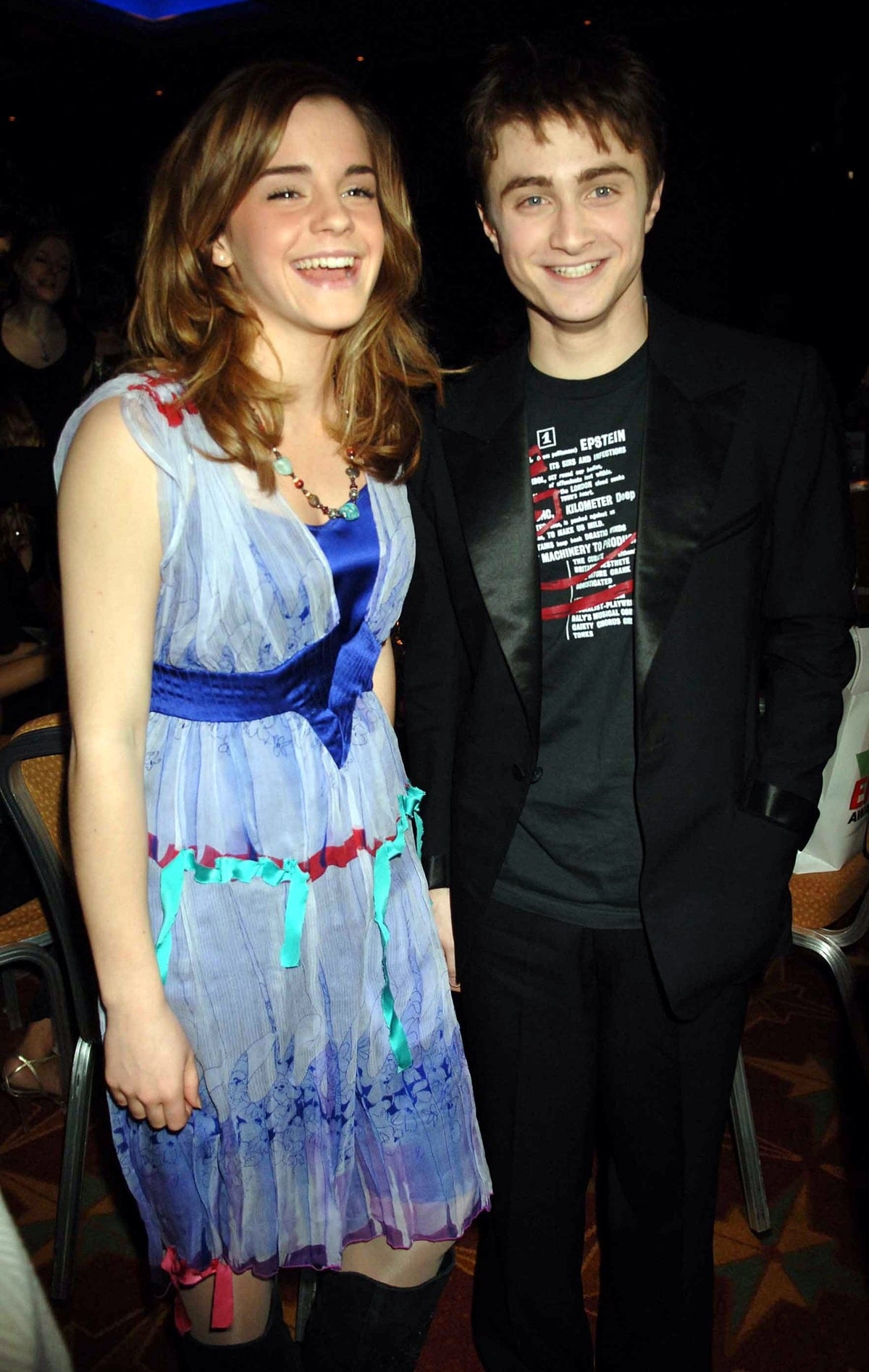 A young Emma Watson and Daniel Radcliffe share a lighthearted moment at an event, with Emma wearing a colorful, whimsical dress and Daniel opting for a casual black blazer and graphic tee ensemble