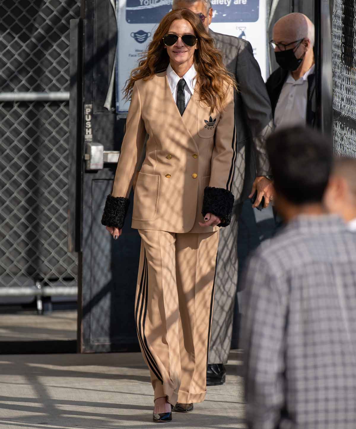 Julia Roberts sports a unique Adidas x Gucci ensemble featuring a beige blazer with black fur cuffs and the Adidas logo on the lapel, paired with matching wide-leg trousers that have the iconic Adidas stripes down the side, and a crisp white shirt with a black tie