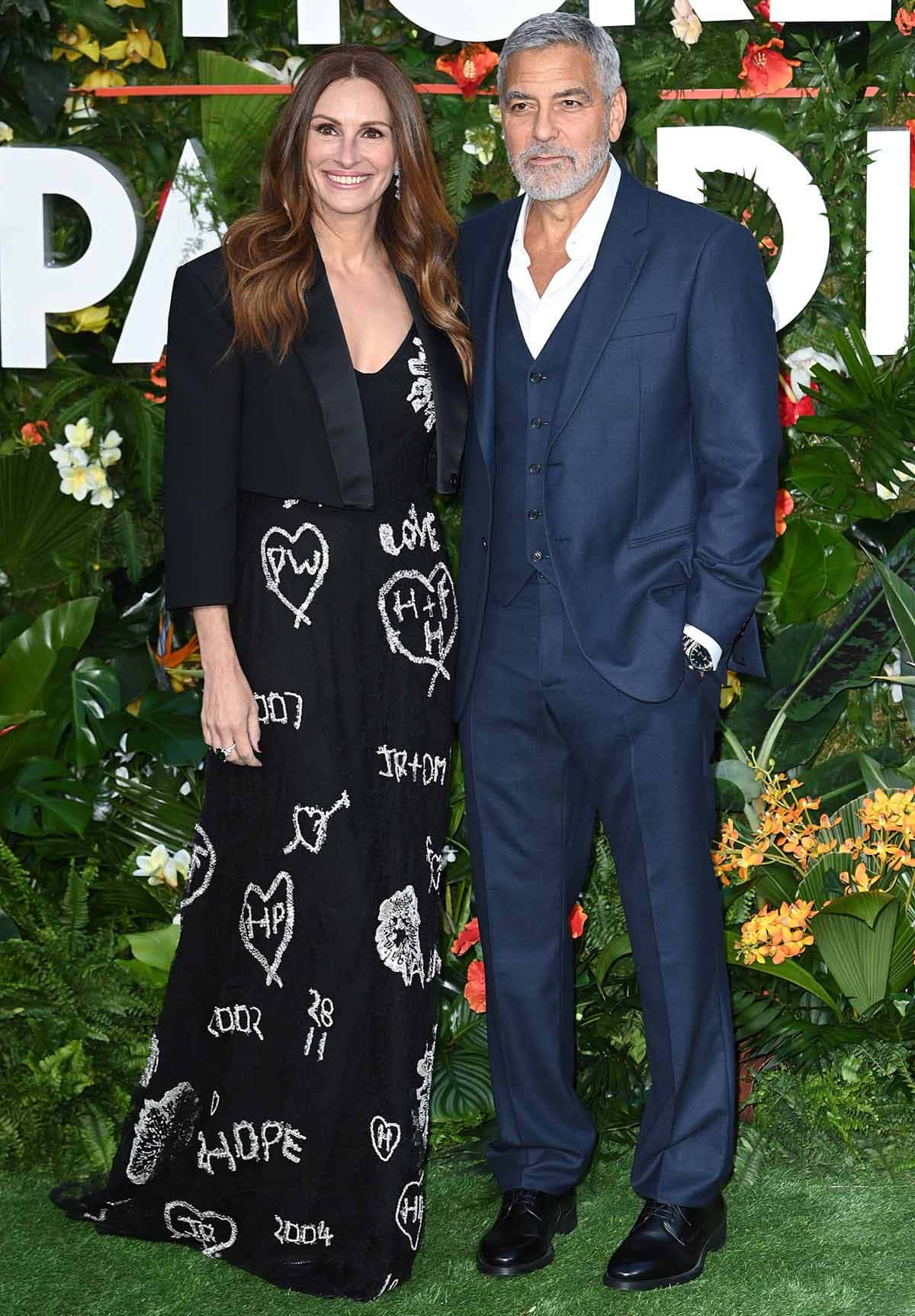 Julia Roberts wears a long black dress with white scribbled designs and a short black blazer, while George Clooney is in a navy blue three-piece suit by Giorgio Armani with a white unbuttoned shirt