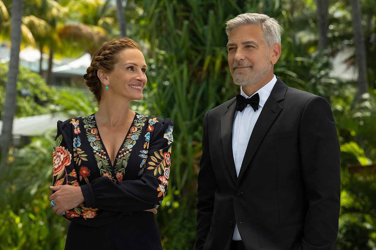 In the romantic comedy Ticket to Paradise, Julia Roberts wears a black Etro Santa Bárbara maxi dress with colorful floral embroidery on the sleeves and neckline, accessorized with turquoise earrings and a matching ring, while George Clooney is dressed in a classic black tuxedo with a bow tie