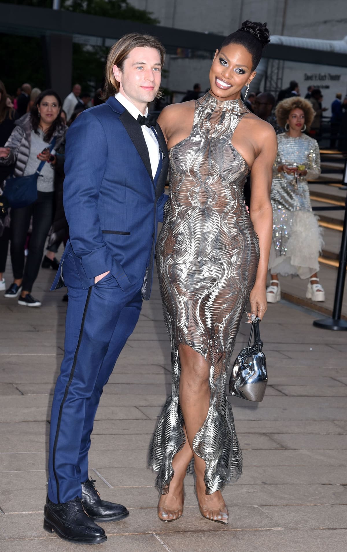 Laverne Cox stuns in a futuristic sheer metallic gown paired with transparent heels, posing alongside Paul McGill at the New York City Ballet's 2022 Fall Fashion Gala