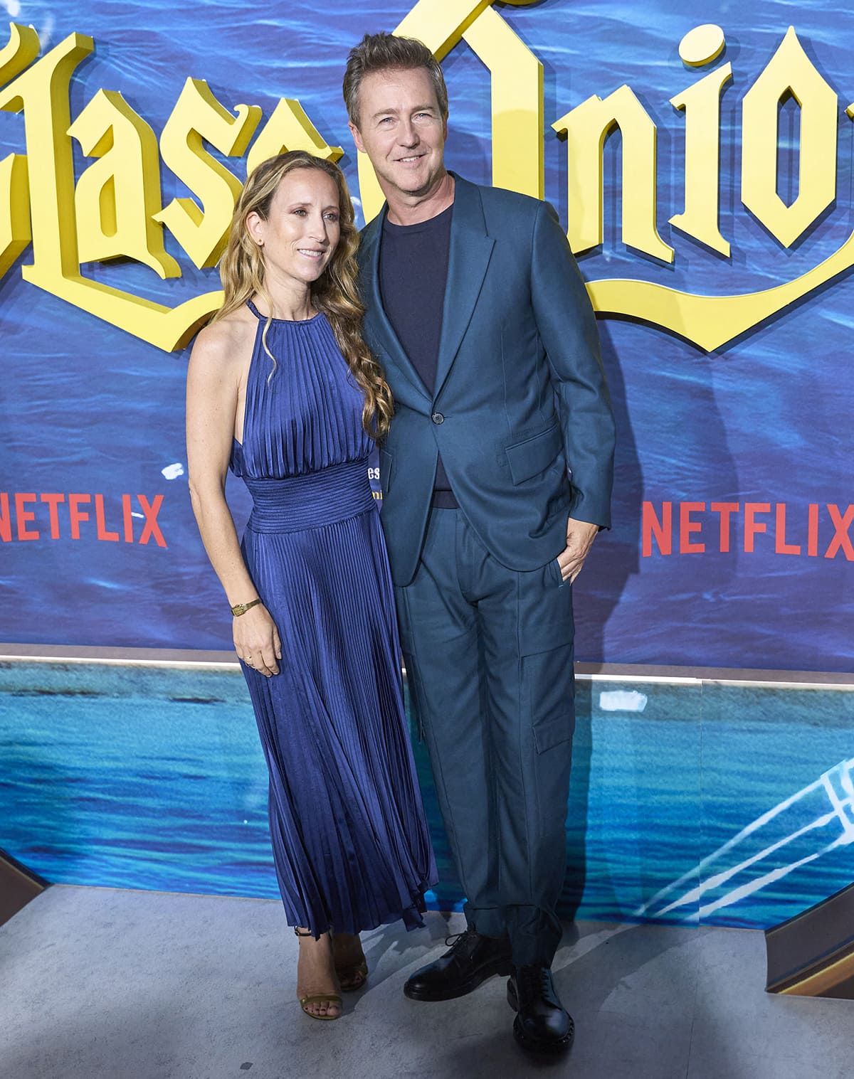 Edward Norton and his wife Shauna Robertson coordinate in matching blue ensembles at the Glass Onion Madrid premiere — Norton in a sleek navy co-ord with black oxford shoes, and Robertson in a pleated halter-neck dress paired with minimalist sandals