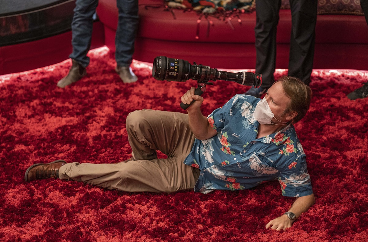 Director Rian Johnson lies on the set of Glass Onion: A Knives Out Mystery, preparing for a shot while holding a camera and wearing a face mask