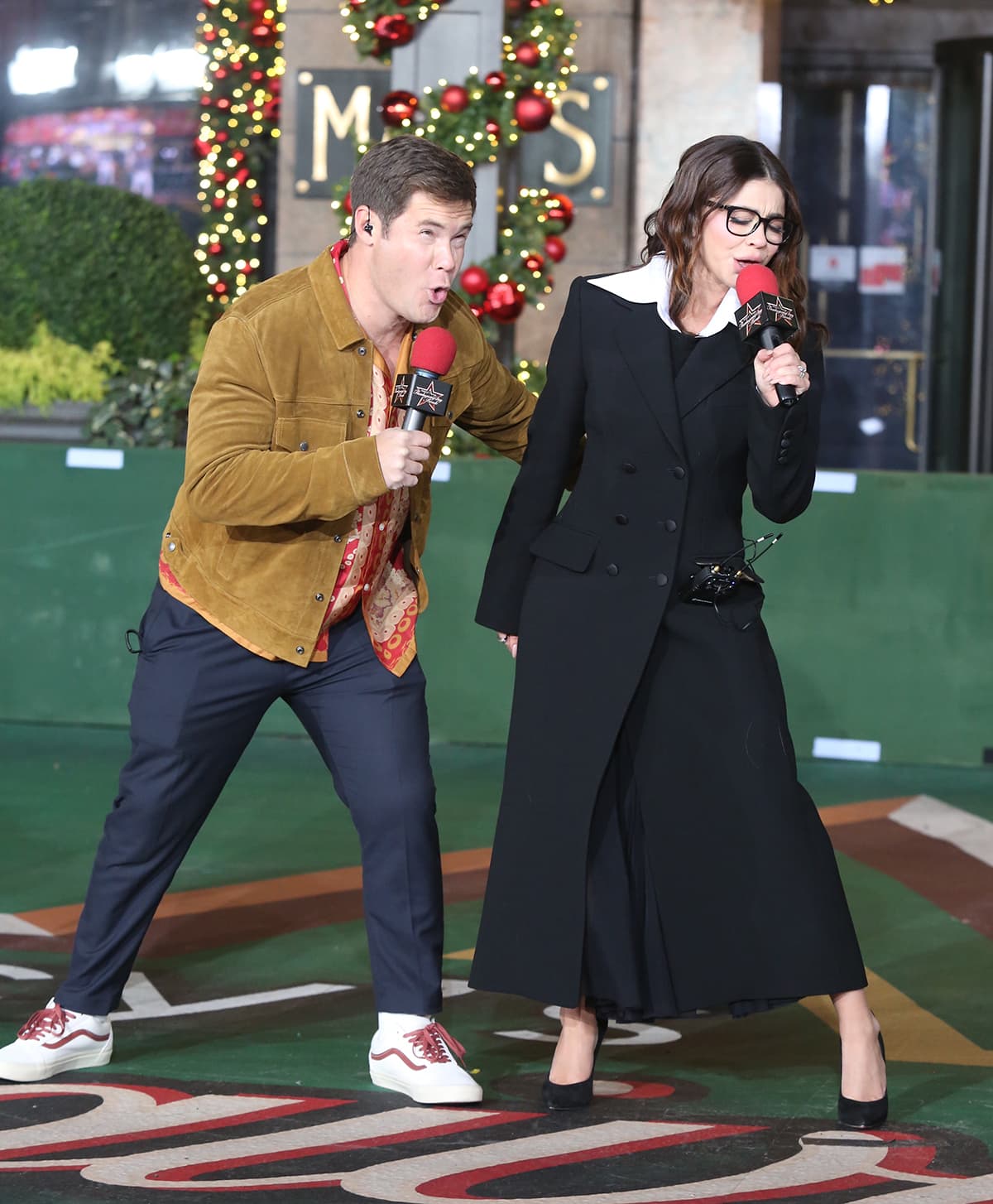Adam Devine keeps things playful in a tan suede jacket, patterned shirt, and white Vans sneakers with red detailing, while Sarah Hyland brings tailored polish to rehearsals in a coat layered over a flowing midi skirt and classic black pumps, as they prepare for the Macy’s Thanksgiving Day Parade on November 22, 2022