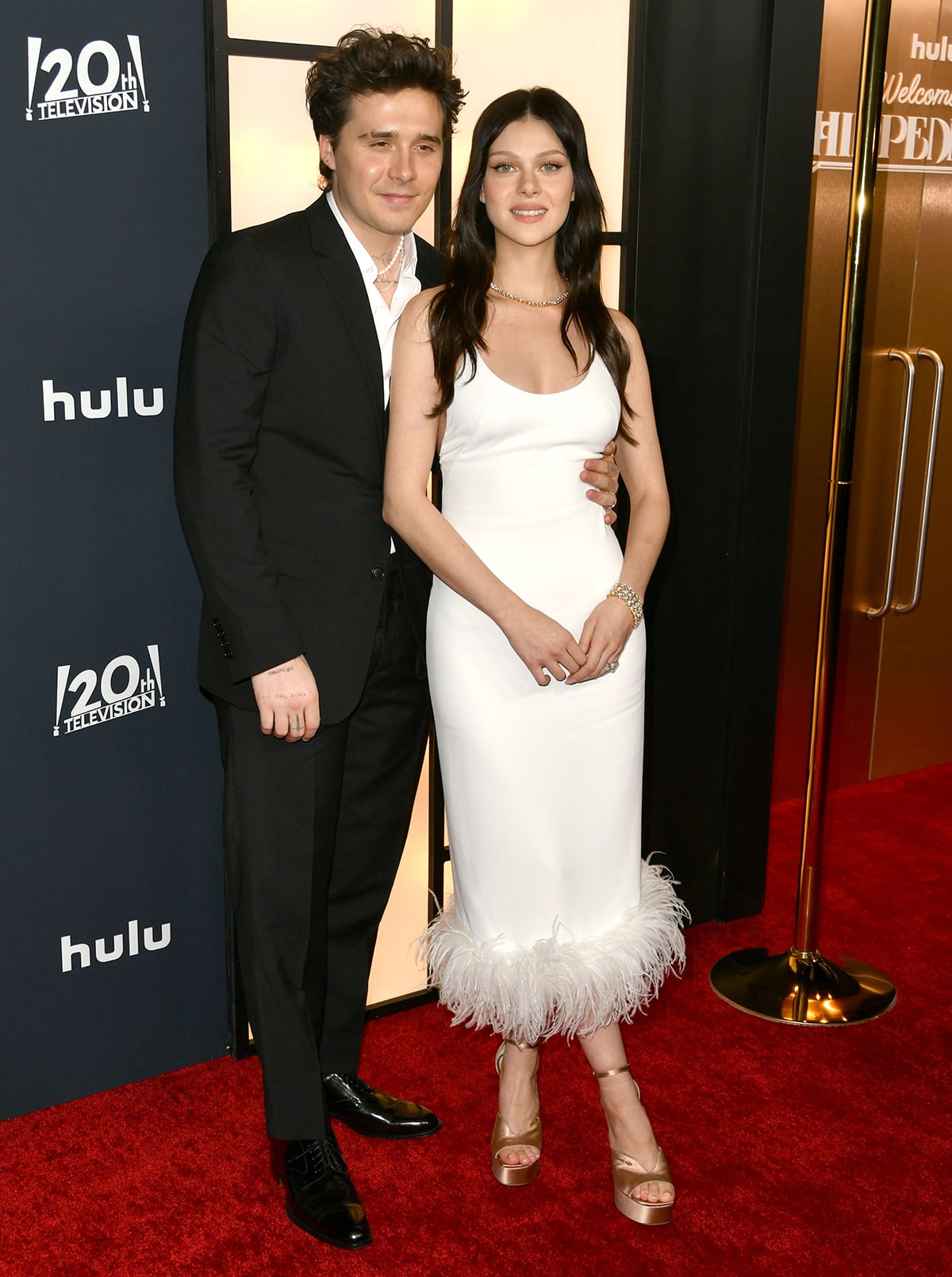 Brooklyn Beckham looks dapper in a black suit with a partially unbuttoned dress shirt and black patent shoes