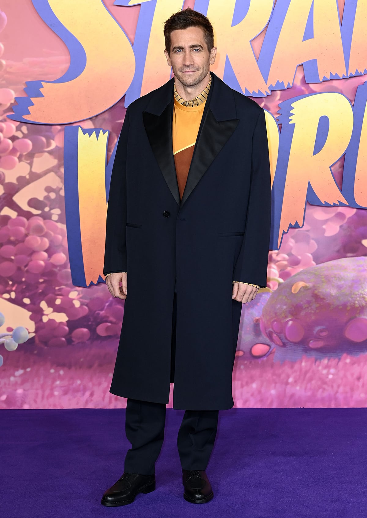 Jake Gyllenhaal cuts a sharp figure in an oversized navy coat and color-blocked sweater, grounding the look with sleek black dress shoes