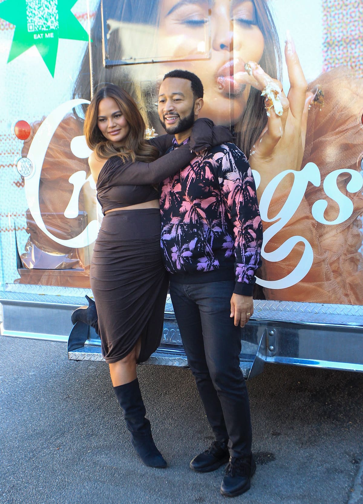 John Legend keeps it casual alongside Chrissy Teigen, sporting a vibrant purple and pink palm tree-printed hoodie paired with black pants and sneakers