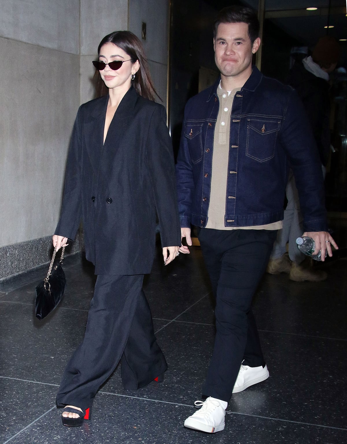 Sarah Hyland sharpens a relaxed, oversized charcoal suit with towering black platform sandals—likely Christian Louboutin, marked by their signature red soles—while Adam Devine keeps things casual in a dark denim jacket, neutral henley, and clean white sneakers as they arrive at the Today Show to promote Bumper in Berlin in New York City on November 23, 2022