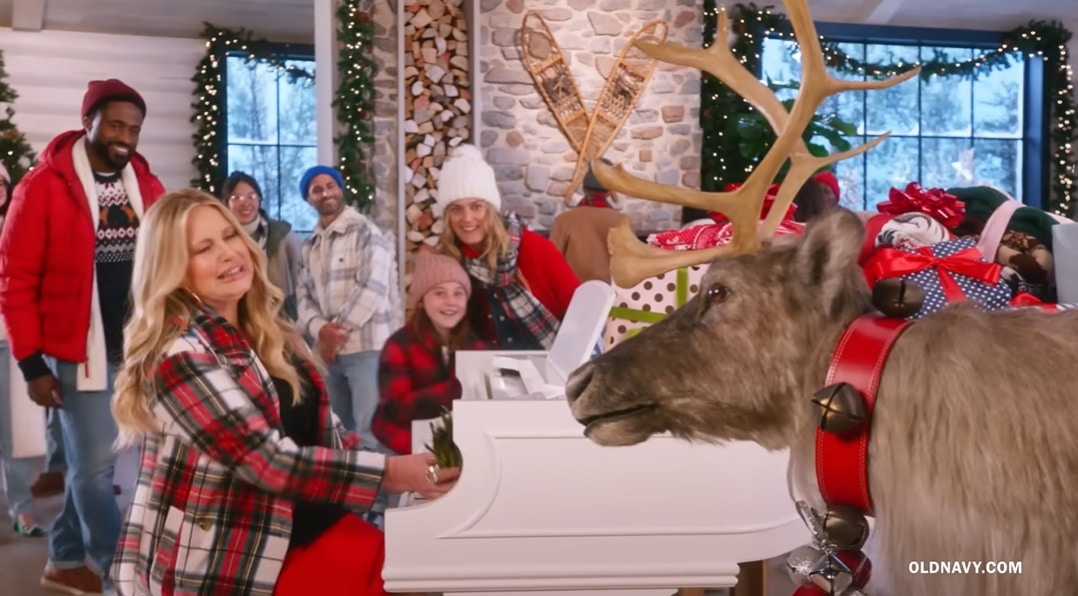 Jennifer Coolidge flashes a festive smile in a red dress and plaid coat while playing the piano beside a reindeer in Old Navy’s “Gifting 2022” holiday commercial — a cozy, tongue-in-cheek scene that perfectly captures her campy holiday charm