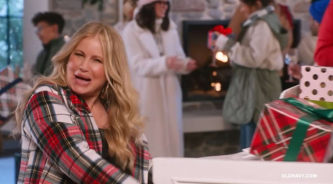 Jennifer Coolidge channels her signature comedic charm while playing the piano in Old Navy’s 2022 holiday commercial, wearing a festive red dress layered with a plaid coat amid a cozy, gift-filled winter scene
