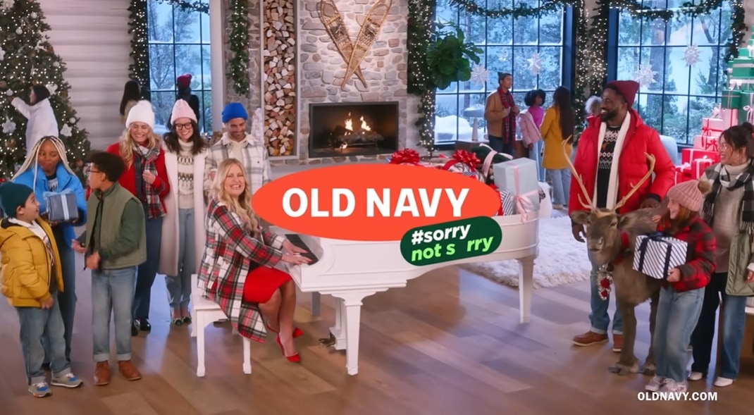Jennifer Coolidge brightens the Old Navy “Gifting 2022” holiday commercial in a festive red dress, plaid coat, and matching red pointed-toe heels while seated at a white piano surrounded by Christmas décor