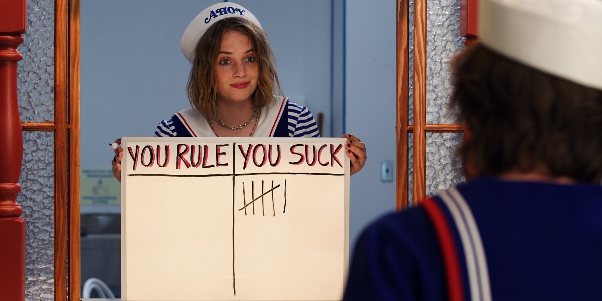 Maya Hawke as Robin Buckley in Stranger Things Season 3, holding her now-iconic “You Rule / You Suck” scoreboard while working at the Scoops Ahoy ice-cream parlor