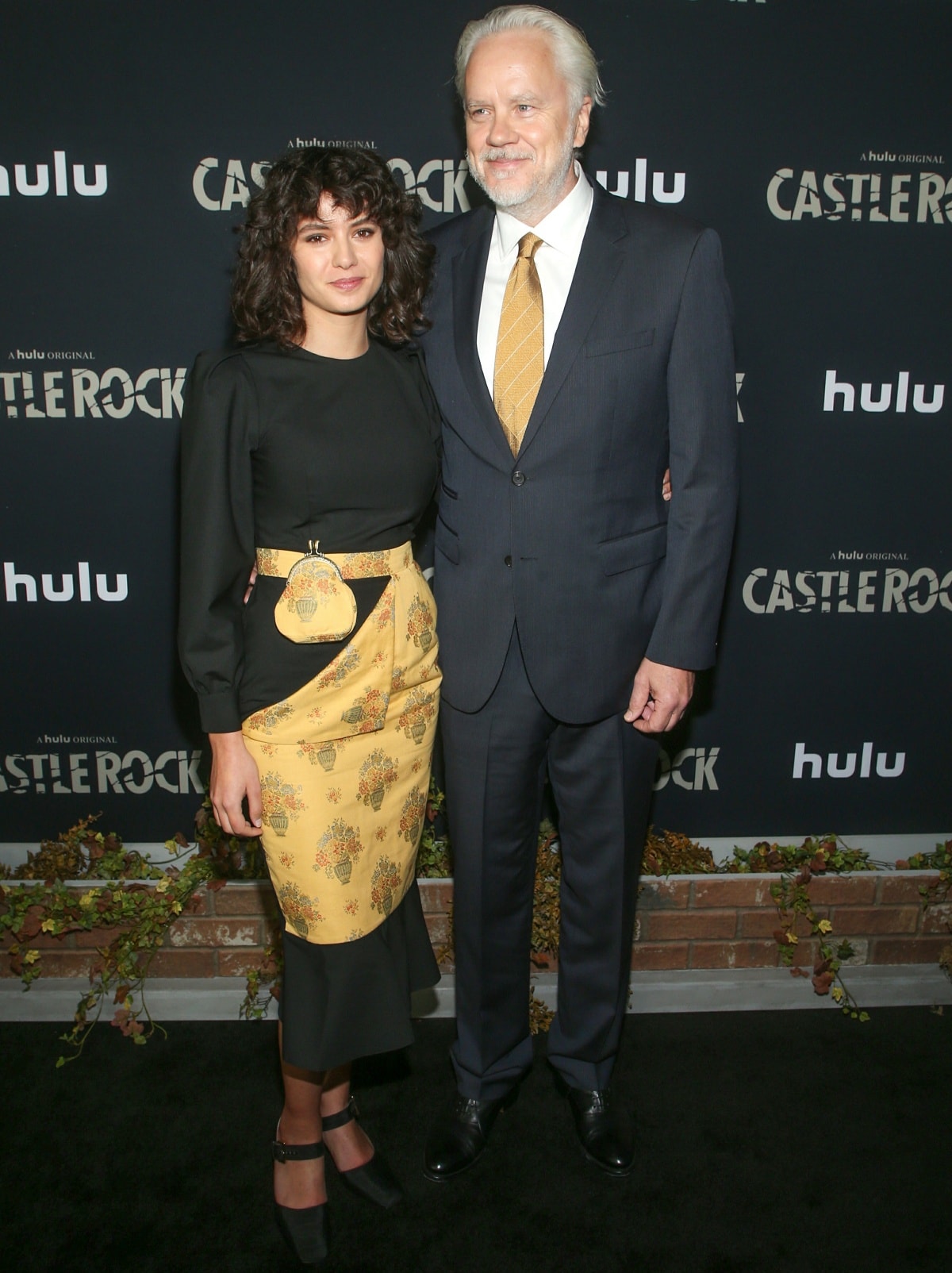 Tim Robbins, standing at 6'4 ¾" (194.9 cm), cut a stately figure in a classic navy suit as he posed with Gratiela Brancusi—radiant in a black-and-gold floral wrap dress—at the Castle Rock Season 2 premiere in Los Angeles on October 14, 2019