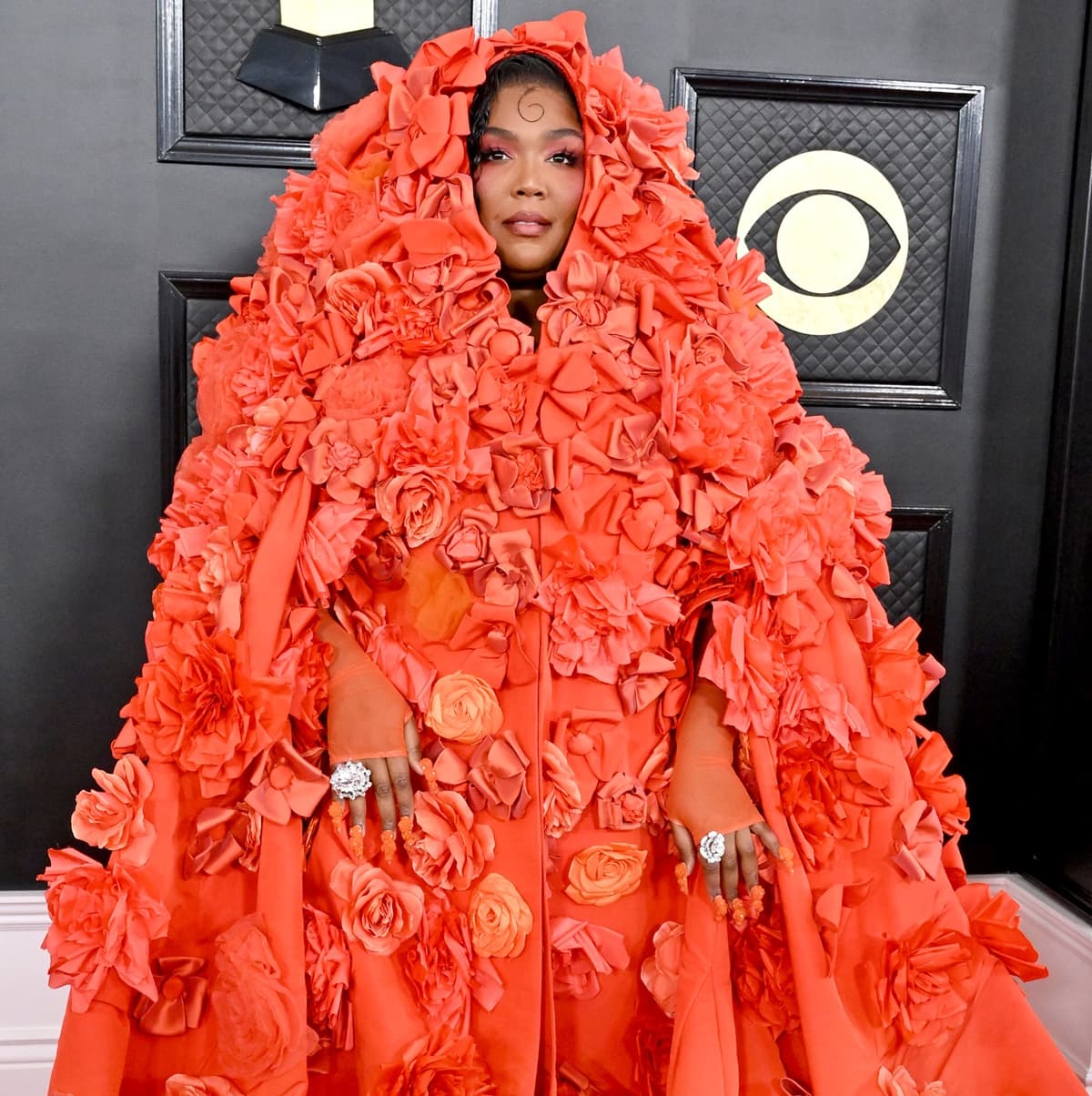 Lizzo framed her face with the oversized hood of her hand-embroidered Dolce Gabbana floral opera coat, finishing the look with diamond flower rings and matching orange nails