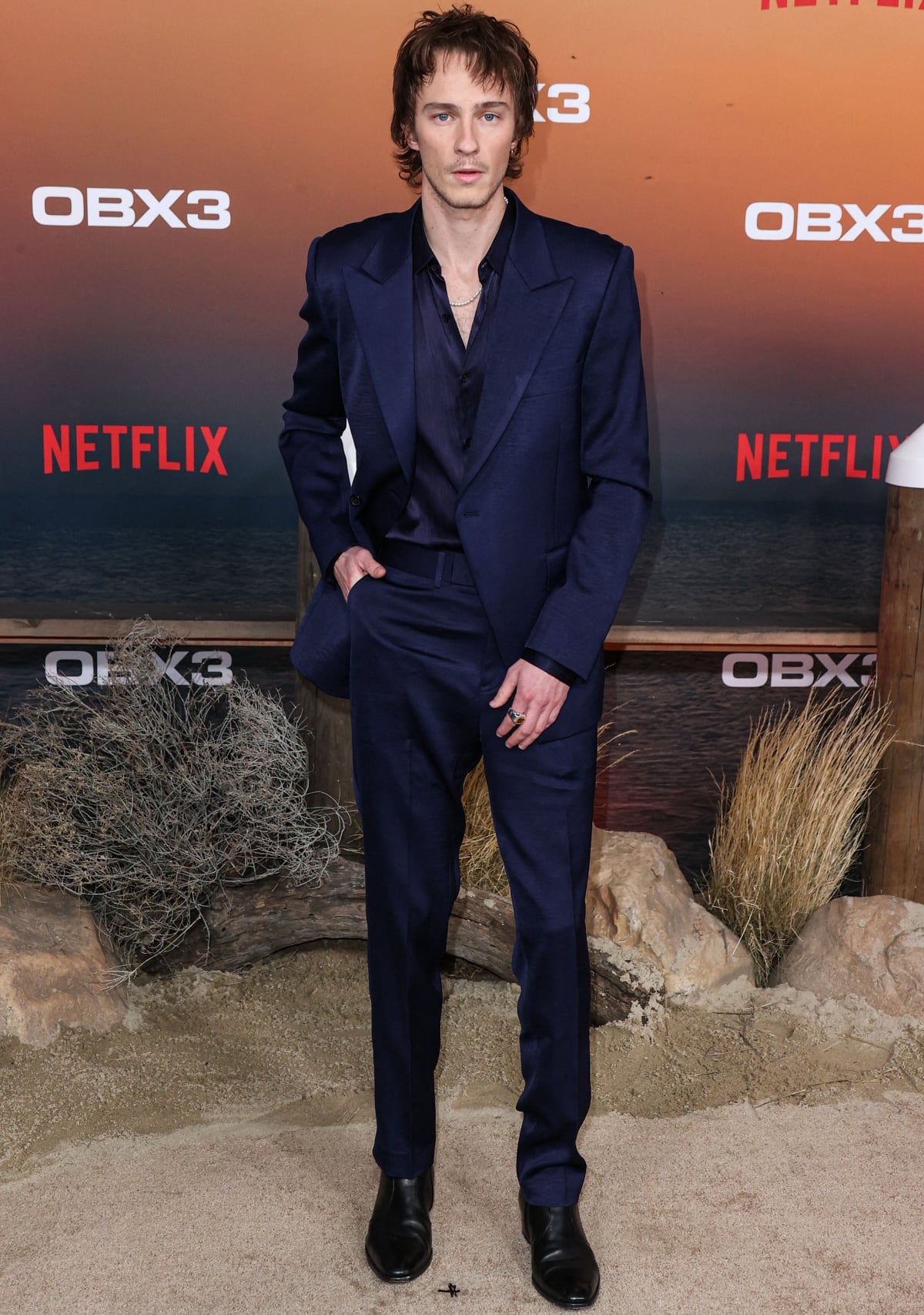 Drew Starkey suited up in a deep navy ensemble with a silky button-down and sharp black leather boots, adding a moody edge to his red carpet look at the premiere of Netflix’s Outer Banks Season 3