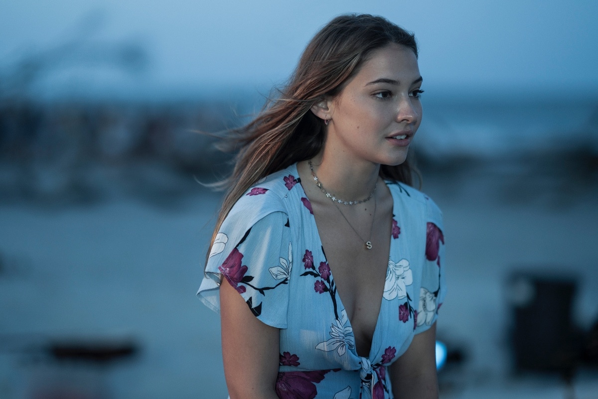 Madelyn Cline wears a plunging blue floral tie-front dress, layered with delicate star and initial necklaces, in a serene beachside scene from Outer Banks