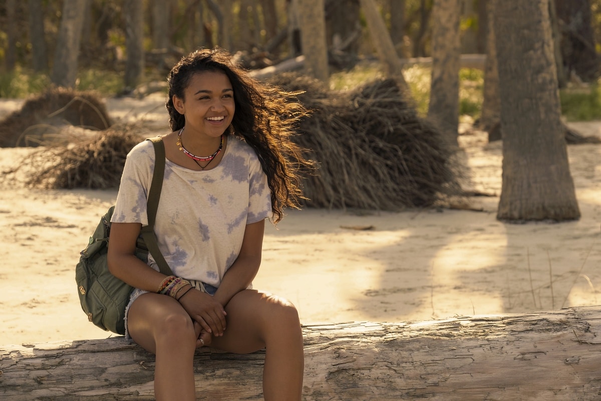 Madison Bailey keeps it casual and beach-ready as Kiara Carrera, wearing a tie-dye tee, cutoff denim shorts, layered beaded necklaces, and a utility backpack in Outer Banks