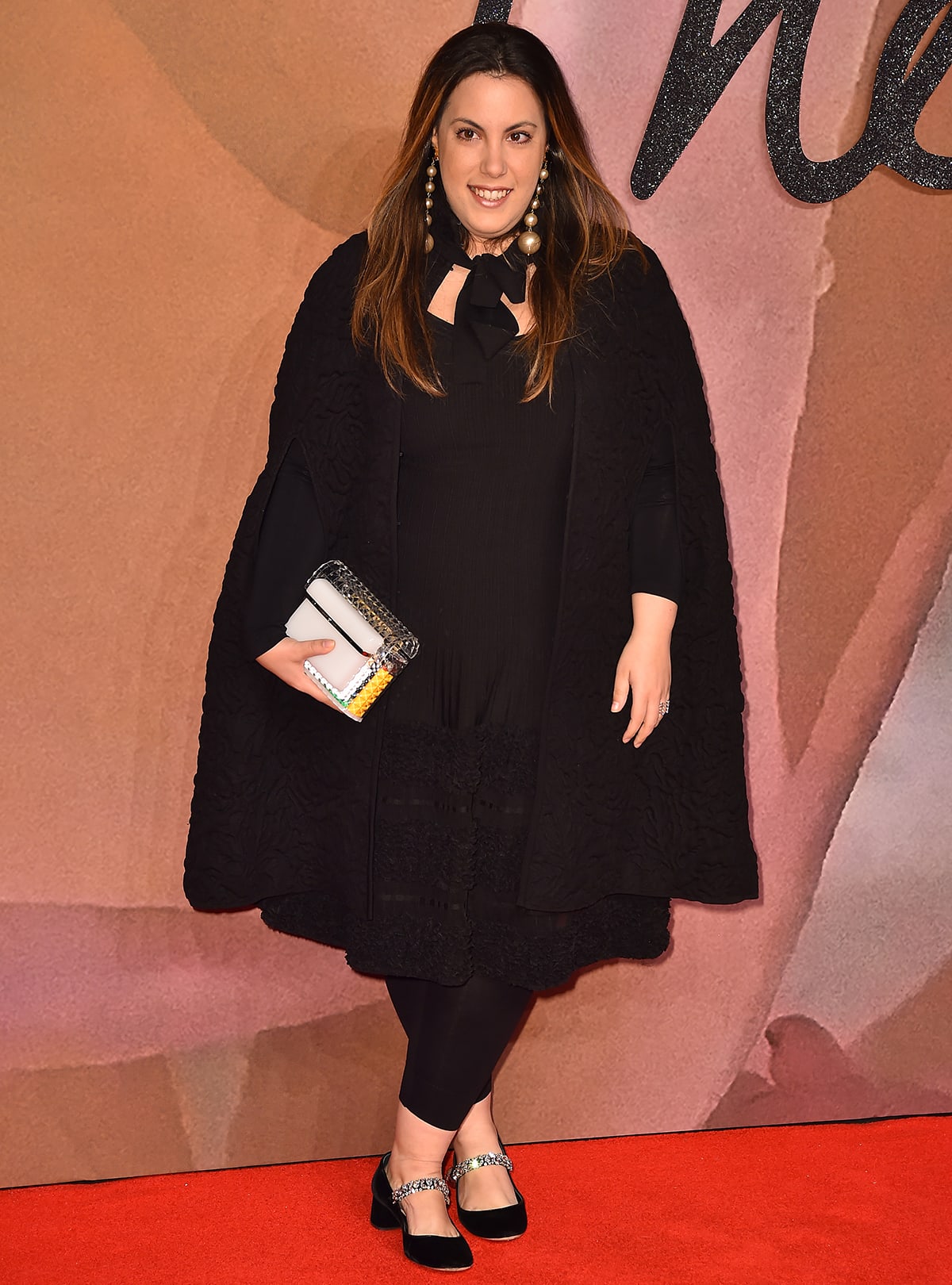 Mary Katrantzou exudes timeless elegance in a textured black ensemble paired with statement earrings, a chic clutch, and embellished Mary Jane heels