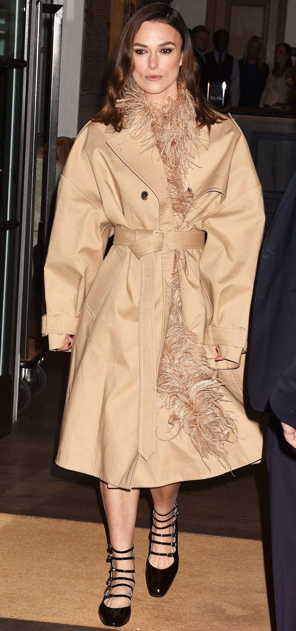 Keira Knightley radiates Old Hollywood glamour in a beige Valentino belted trench coat trimmed with tonal feathers, paired with black velvet Carel Paris “Xena” pumps featuring multiple buckle straps and low block heels