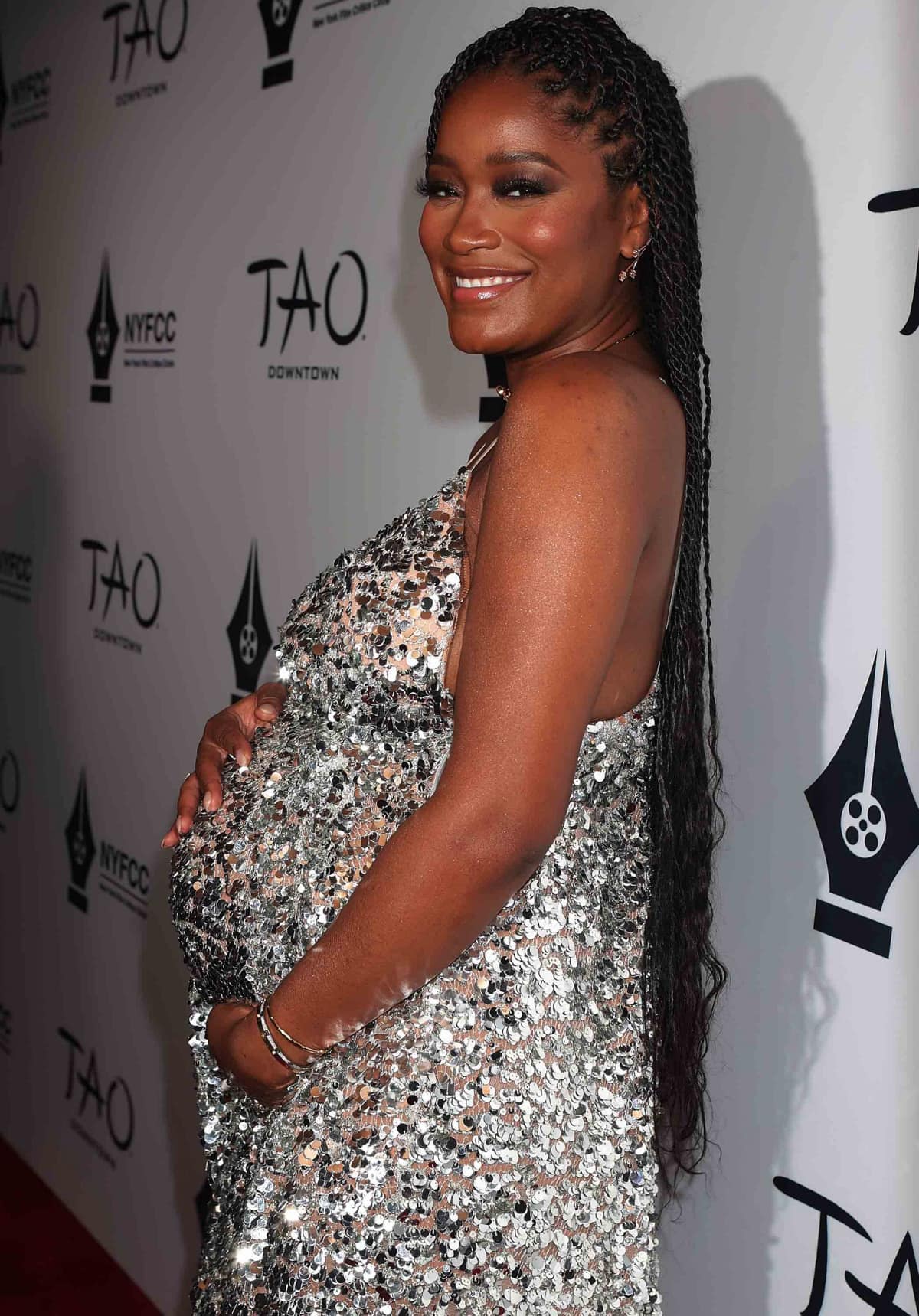 Keke Palmer showcases her baby bump in a silver sequined Michael Kors gown, styling the look with long twisted braids, delicate stud earrings, and sleek metallic bangles