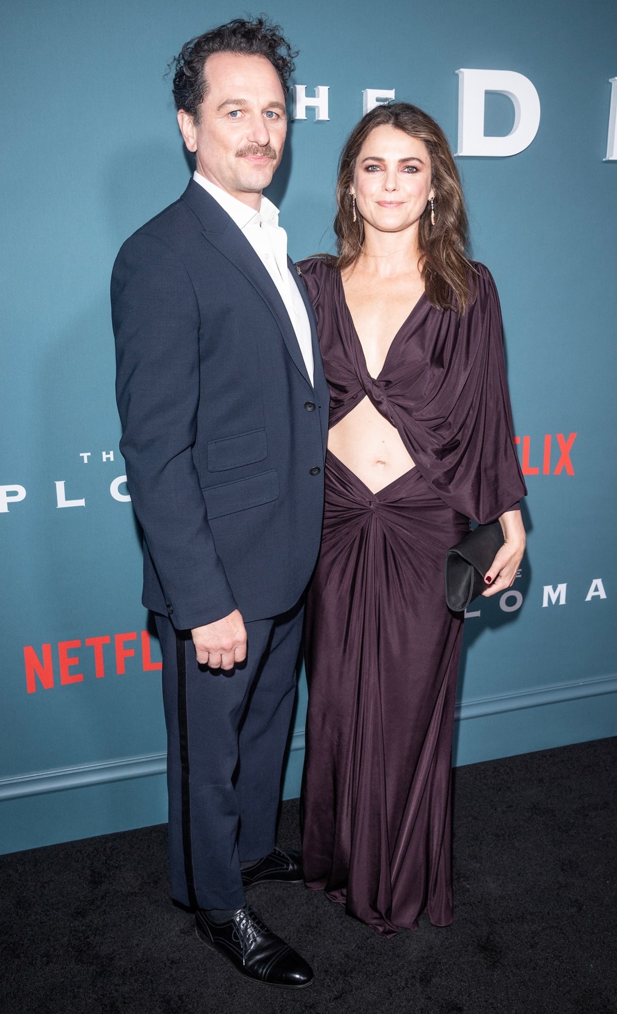 Matthew Rhys, looking sharp in a navy tuxedo with satin side stripes and black cap-toe dress shoes, posed alongside his wife Keri Russell, who stunned in a plum Saint Laurent cutout gown and strappy heels mostly hidden beneath the floor-length hem