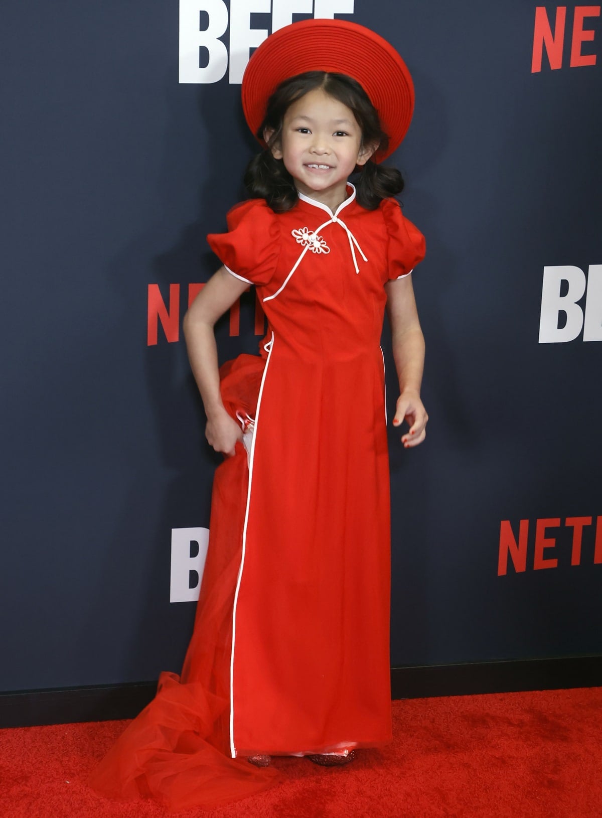Remy Holt lit up the Beef red carpet in an adorable red puff-sleeve dress with white piping, a matching wide-brimmed hat, and sparkly shoes peeking out from beneath her tulle train