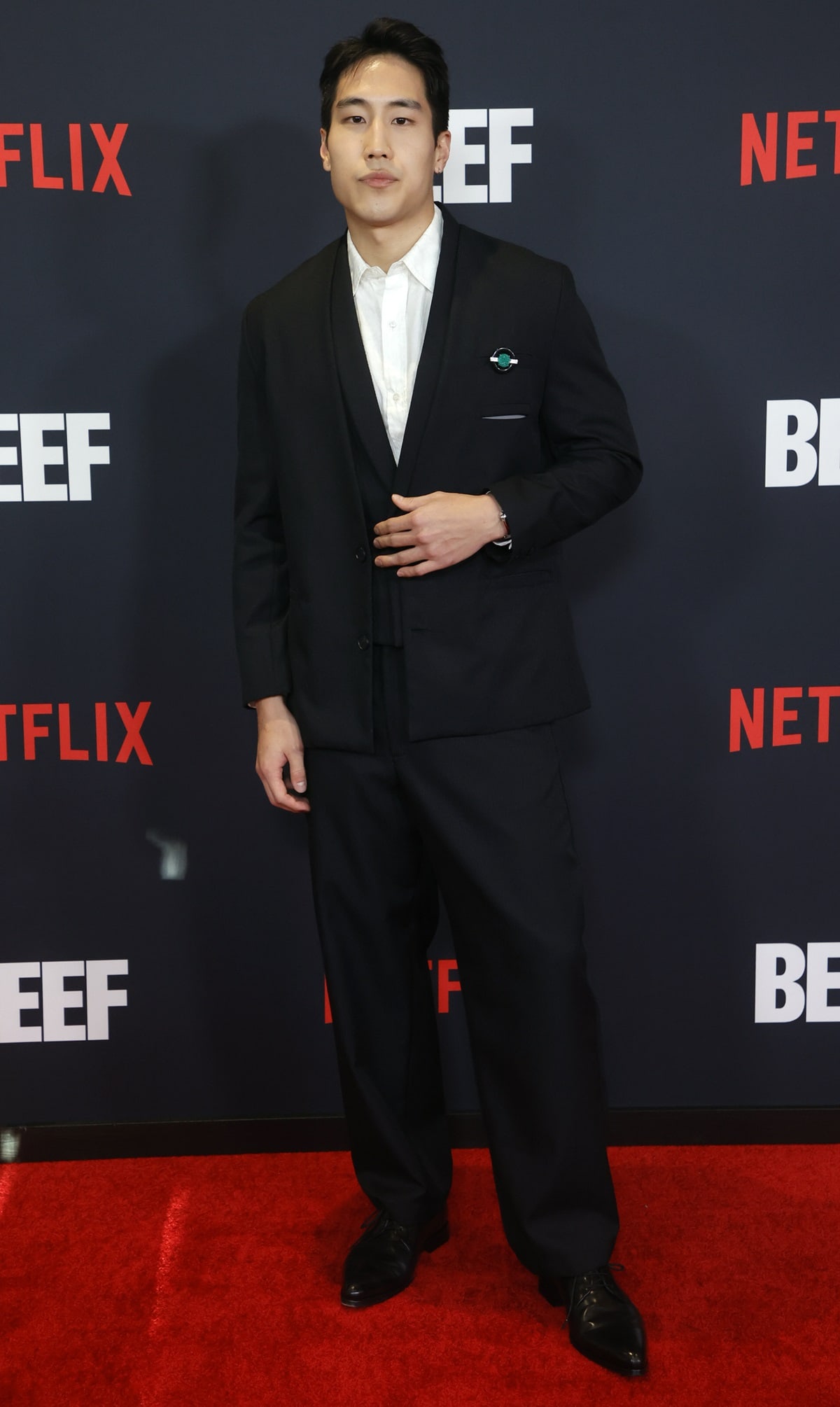 Young Mazino looked sharp at the Beef premiere in a classic black suit with a crisp white shirt, pointed dress shoes, and a statement emerald brooch for a touch of flair