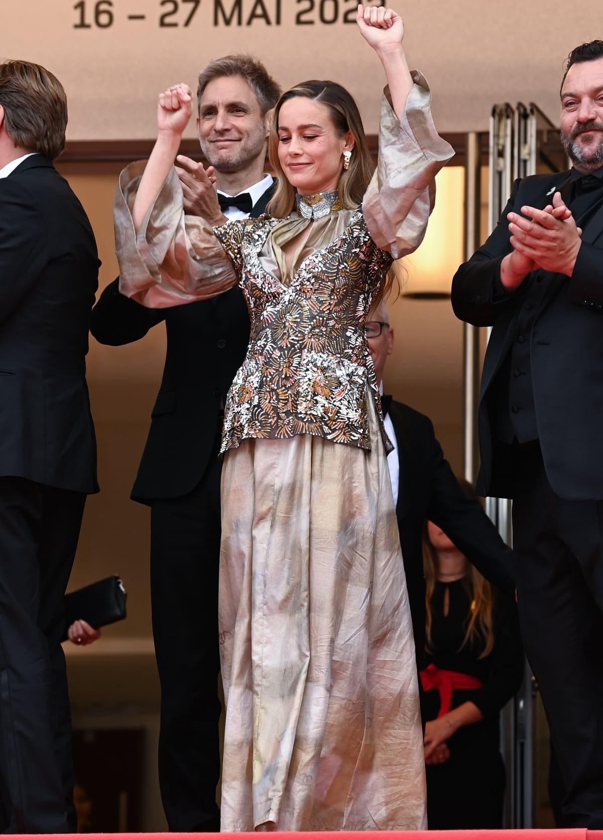 Brie Larson raises her arms in celebration while wearing a Chanel Resort 2024 gown with voluminous sleeves, a sequined bodice, and a softly crinkled gold-beige skirt, accessorized with drop earrings and a statement choker at the 76th Cannes Film Festival